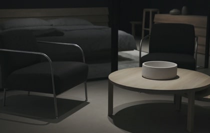 A modern, minimalist interior setting with a light-colored wooden coffee table in the foreground. The table has a white bowl on it, surrounded by two black armchairs with metal armrests. In the background, there's a bed with a dark-colored bedspread and pillows, and a wooden headboard.