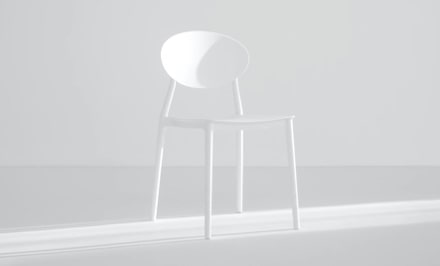 a modern white chair in a plain, light gray setting. The chair has a round back and thin legs, blending in with the simple background. Soft shadows to the right add a bit of contrast, highlighting the chair’s minimalist design.